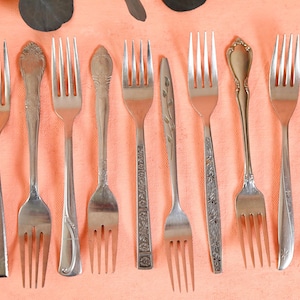 May include: A collection of ten silver forks with varying designs. The forks are arranged in a row on a peach-colored surface.