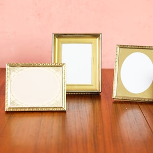 May include: A collection of vintage picture frames in various shapes and sizes. The frames are gold and bronze, with oval and rectangular openings. They are arranged on a wooden surface against a pink background.