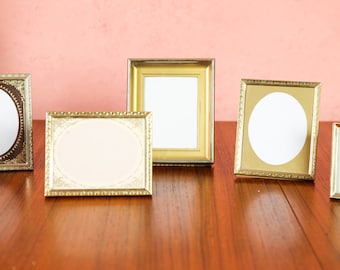May include: A collection of vintage picture frames in various shapes and sizes. The frames are gold and bronze, with oval and rectangular openings. They are arranged on a wooden surface against a pink background.