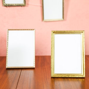 May include: Five ornate gold picture frames with white mats, four are on a wooden table and one is on a pink wall.