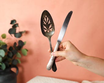 May include: A silver cake server and a silver butter knife are held in a hand against a peach colored background.