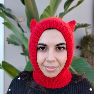 The image features a young woman wearing a red balaclava with cat ears. The balaclava covers ears, throat and neck.