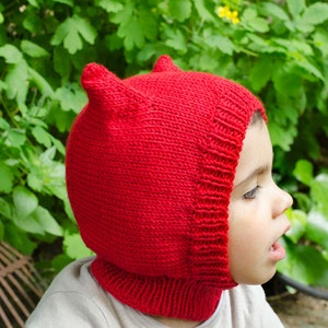 May include: A child wearing a red knitted hat with ear flaps. The hat is made with a simple stitch pattern and has a pointed top.