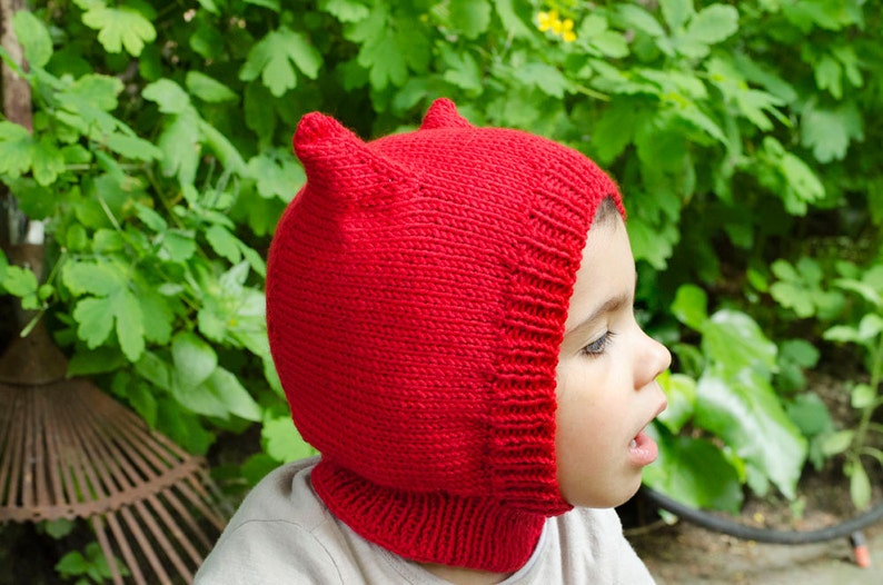 May include: A child wearing a red knitted hat with ear flaps. The hat is made with a simple stitch pattern and has a pointed top.