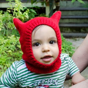May include: A child wearing a red knitted hat with horns and a green and white striped shirt with the text "Super Team" printed on it.