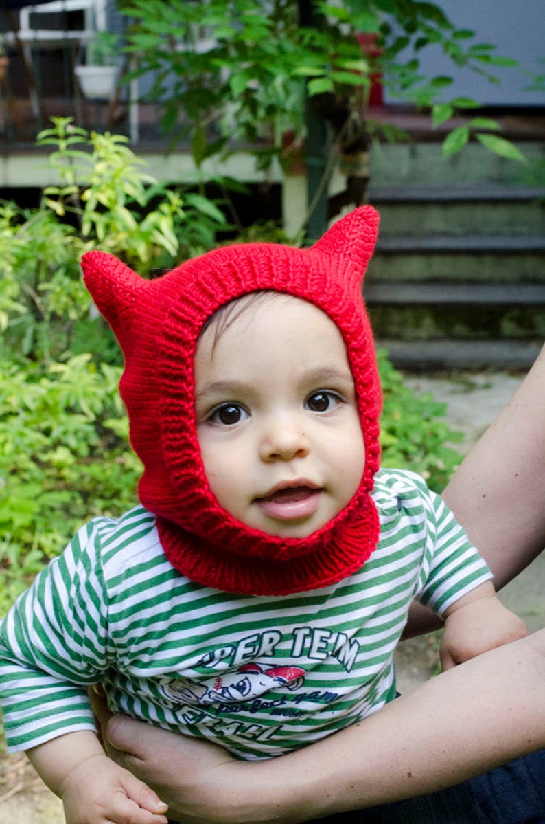 May include: A child wearing a red knitted hat with horns and a green and white striped shirt with the text "Super Team" printed on it.