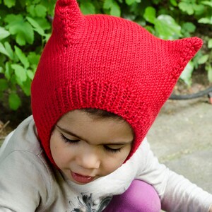 May include: A child wearing a red knitted hat with cat ears. The hat is pulled down over the child's head, covering their ears and forehead. The child is looking down at something out of frame.