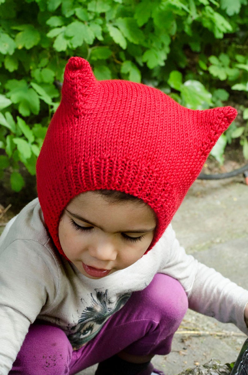 May include: A child wearing a red knitted hat with cat ears. The hat is pulled down over the child's head, covering their ears and forehead. The child is looking down at something out of frame.