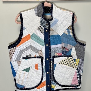 May include: A quilted vest with a patchwork design in various colors, including white, blue, orange, and plaid patterns. The vest has a denim-like front closure with snap buttons and two front pockets. The edges are trimmed with a dark blue fabric.
