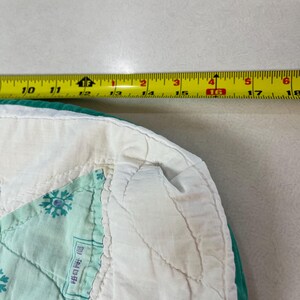 May include: A quilted fabric piece with a white border and a teal edge, featuring a light green snowflake pattern and a red and white checkered section. A yellow measuring tape is positioned above, showing a width of approximately 8 inches.
