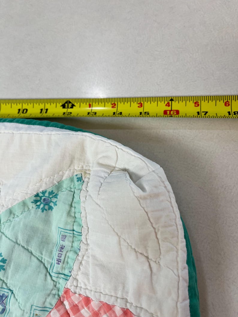 May include: A quilted fabric piece with a white border and a teal edge, featuring a light green snowflake pattern and a red and white checkered section. A yellow measuring tape is positioned above, showing a width of approximately 8 inches.