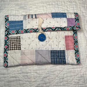 May include: A handmade patchwork clutch with a button closure. The fabric features a variety of patterns, including blue, pink, and brown squares, and is bordered with a floral design. The button is a bright blue color.