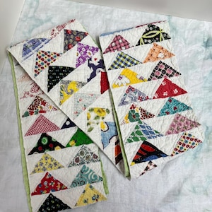May include: Two vintage handmade quilts with a patchwork design. Each quilt features a white background with colorful triangular patches in various patterns and colors, including floral, geometric, and solid hues. The edges are finished with a green binding.