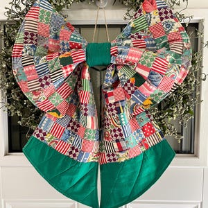 May include: A large decorative bow constructed from a patchwork of various colorful fabric squares featuring stripes, floral patterns, and gingham prints. The center and the bottom tails of the bow are made of solid green fabric.