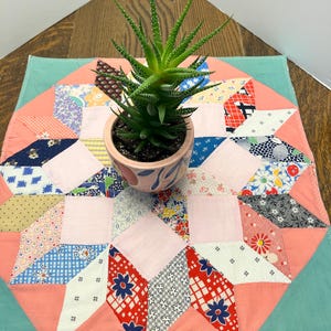 May include: A square fabric mat featuring a multi-colored patchwork star pattern in shades of pink, blue, red, and white, topped with a green succulent plant in a small, round, light pink ceramic pot with blue leaf-shaped painted accents.