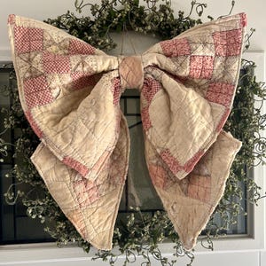 May include: A large decorative bow constructed from quilted fabric featuring a patchwork pattern of cream, off-white, and faded red checkered squares. The fabric has a visible stitched texture throughout, forming a soft, dimensional bow shape.