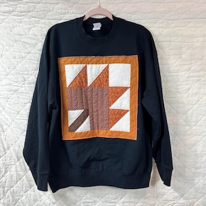 May include: A black sweatshirt featuring a quilted square design. The square has a brown, white, and orange star pattern. The sweatshirt is hanging on a pink hanger against a white background.