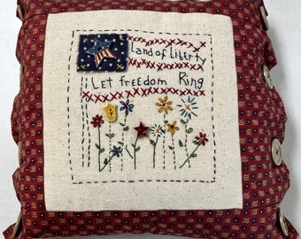 May include: A square fabric pillow featuring a cream-colored center panel with embroidered flowers, a blue star patch, and the text "Land of Liberty" and "Let Freedom Ring." The border is red fabric with a small pattern and wooden buttons on the sides.