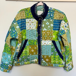 May include: A patchwork jacket in shades of green, blue, and olive green, featuring various floral and geometric patterns. The jacket has a navy blue corduroy collar and trim, with a button-up front and two front pockets.