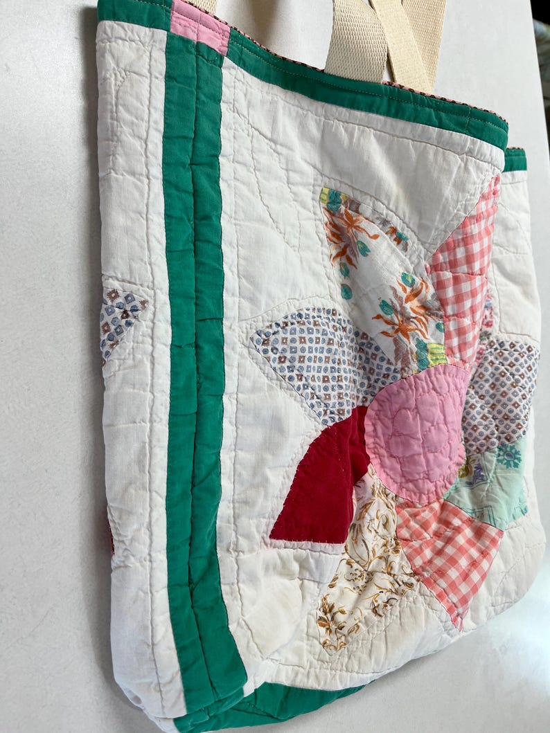 May include: A quilted tote bag featuring a patchwork floral design in shades of pink, red, and white with green fabric borders. The bag is constructed from various patterned fabric pieces and includes two beige fabric handles at the top.