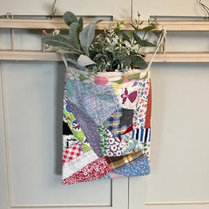 May include: A patchwork fabric hanging planter filled with faux greenery. The planter is made of various patterned fabrics in red, blue, and white. The planter hangs from a white wooden frame.