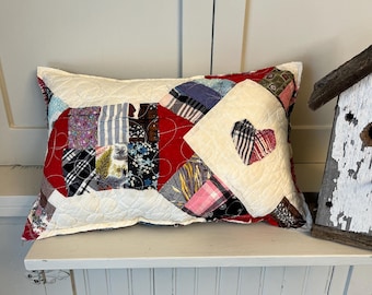 May include: A decorative pillow with a patchwork design. The pillow features a variety of fabric patterns in red, white, blue, and floral prints. A heart-shaped cutout reveals a striped pattern. The pillow is placed on a white shelf.