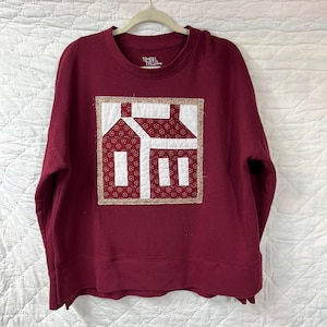 May include: A burgundy sweatshirt with a crew neck and long sleeves. The front features a square patch with a house design in white and red fabric, framed by a gold border. The brand name "TIME TRU" is visible on the neckline.