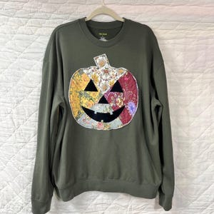 May include: Olive green sweatshirt with a patchwork pumpkin design. The pumpkin features floral and patterned fabric in yellow, red, and white. The pumpkin has black triangle eyes and a curved black mouth. The sweatshirt is hanging on a hanger.