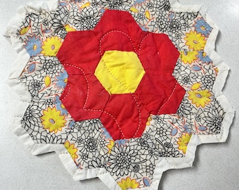 May include: A hexagonal patchwork quilt coaster featuring a central yellow hexagon surrounded by six red petals. The outer border consists of smaller hexagons with black and white floral line art, yellow flowers, and light blue accents.