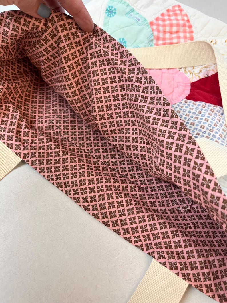 May include: A tote bag featuring a pink and brown geometric patterned fabric lining. The exterior is constructed from a patchwork quilt design with various colors and textures, including red velvet and light blue fabric, with cream-colored straps.