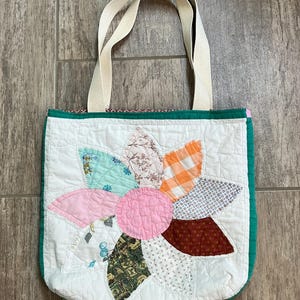 May include: A quilted tote bag featuring a large, multi-colored flower appliqu&eacute; on a white background. The flower petals are made from various patterned fabrics in shades of pink, orange, blue, green, and brown. It has two cream-colored fabric handles.