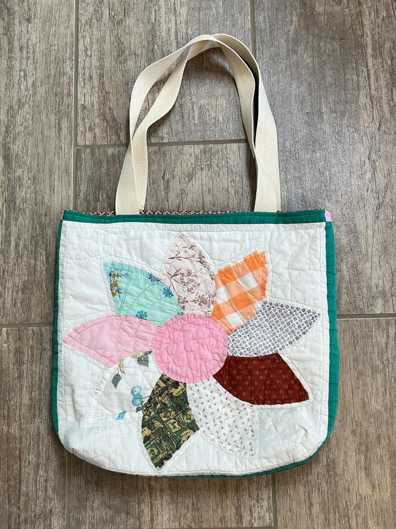May include: A quilted tote bag featuring a large, multi-colored flower appliqu&eacute; on a white background. The flower petals are made from various patterned fabrics in shades of pink, orange, blue, green, and brown. It has two cream-colored fabric handles.