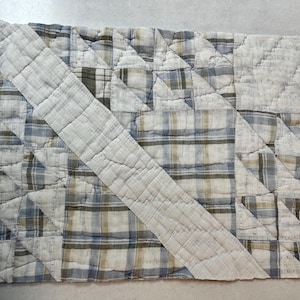 May include: A rectangular quilted placemat featuring a patchwork design of blue, tan, and white plaid fabric squares. The piece is constructed with visible stitching and a prominent diagonal band of solid white fabric running across the surface.