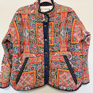 May include: A colorful quilted jacket with a vibrant floral and geometric pattern in red, yellow, blue, and green. The jacket features a denim-like trim and brass-colored buttons. The jacket is hanging on a hanger.
