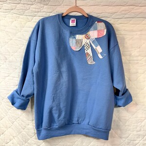 May include: A light blue sweatshirt with a patchwork bow applique on the front. The bow features various patterned fabrics in shades of red, blue, and white. The sweatshirt has a crew neck and long sleeves.