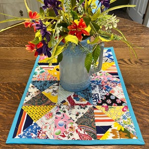May include: A light blue pitcher filled with colorful flowers sits on a patchwork table runner. The runner features a variety of patterned fabrics in triangles, bordered by a bright blue edge. The table is made of wood.