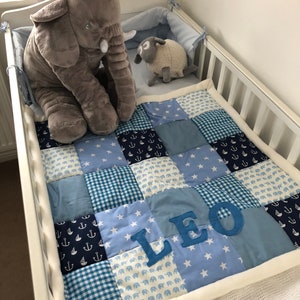 May include: A cot with a patchwork quilt in shades of blue, patterned with stars, anchors, and elephants, with the name "LEO". A grey elephant and a sheep soft toy are placed in the cot. The quilt is approximately 100cm x 75cm.