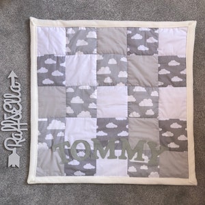 May include: A grey and white patchwork baby quilt with a white border. The quilt has a cloud pattern and the name "TOMMY" is spelled out in grey letters.