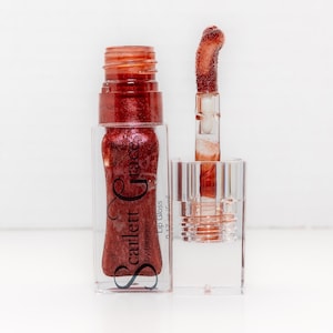 May include: A clear lip gloss tube with a red, glittery liquid and a matching applicator. The label reads "Scarlett Grace" in a cursive font. The applicator is dipped in the gloss, showcasing its color and texture. The product is set against a white background.