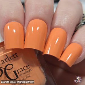 May include: Close-up of a hand with orange nail polish. The polish is a creamy, opaque orange color. The nail polish bottle is partially visible, with the text "Scarlett Grace- Cosmos & Cream". The polish is vegan and cruelty-free.