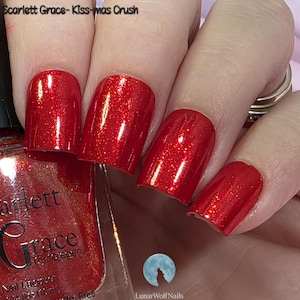 May include: Close-up of red nail polish with glitter, applied to fingernails. The nail polish bottle is visible, with the brand name "Scarlett Grace" and the text "Kiss-mas Crush". The nails are a vibrant, festive red.