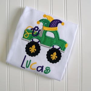May include: A white t-shirt with an embroidered green monster truck with purple and yellow jester hats. The truck has black wheels with gold fleur-de-lis. The name "Lucas" is embroidered in yellow and purple below the truck.