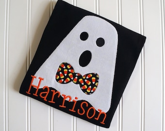 May include: Black t-shirt with a white ghost applique wearing a candy corn bow tie. The shirt is personalized with the name "Harrison".