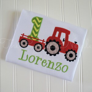 May include: A white onesie with a red tractor pulling a green and yellow wagon. The onesie is embroidered with the name "Lorenzo".