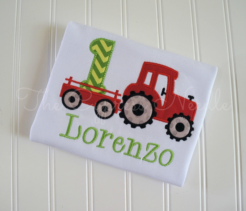 May include: A white onesie with a red tractor pulling a green and yellow wagon. The onesie is embroidered with the name "Lorenzo".