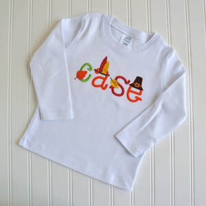 Puede incluir: Camiseta blanca de manga larga con el nombre "Case" bordado en naranja, verde y marr&oacute;n con detalles de oto&ntilde;o, incluyendo un sombrero de peregrino, hojas y una pluma de pavo.