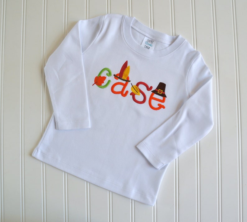 Puede incluir: Camiseta blanca de manga larga con el nombre "Case" bordado en naranja, verde y marr&oacute;n con detalles de oto&ntilde;o, incluyendo un sombrero de peregrino, hojas y una pluma de pavo.