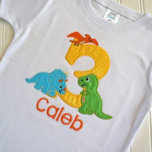 May include: White t-shirt with a yellow number 3, a green dinosaur, a blue dinosaur, and an orange dinosaur. The name Caleb is written in orange below the number.