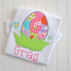 May include: White baby onesie with a colorful patchwork Easter egg design. The egg has a pink, purple, green, and yellow pattern. The onesie has the name "Gray" embroidered in pink below the egg.