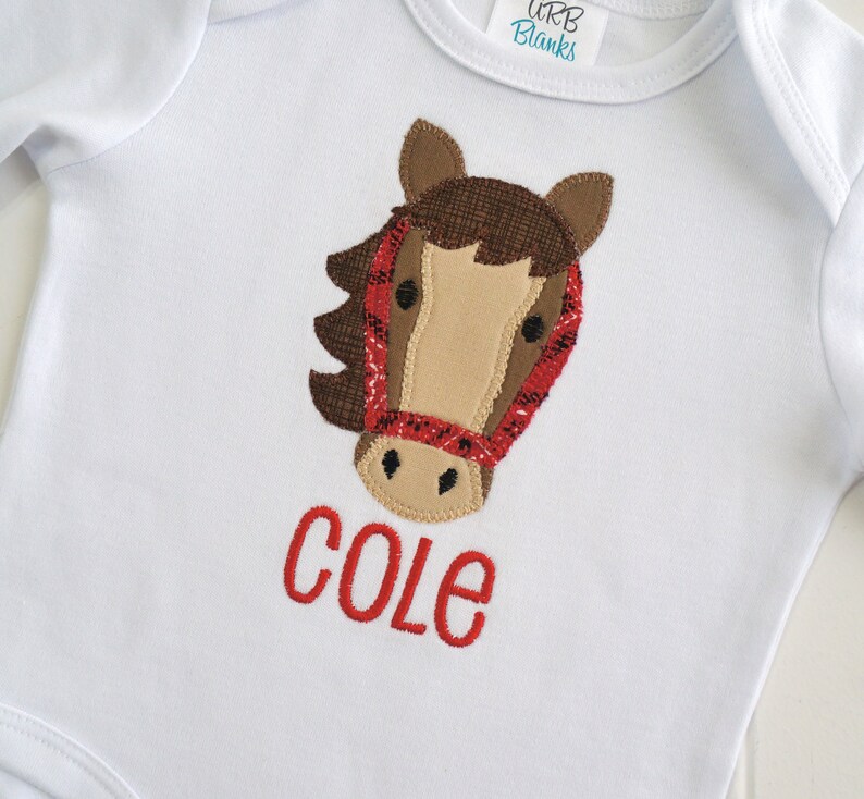May include: White baby onesie with a brown and red horse applique and the name "COLE" embroidered in red.